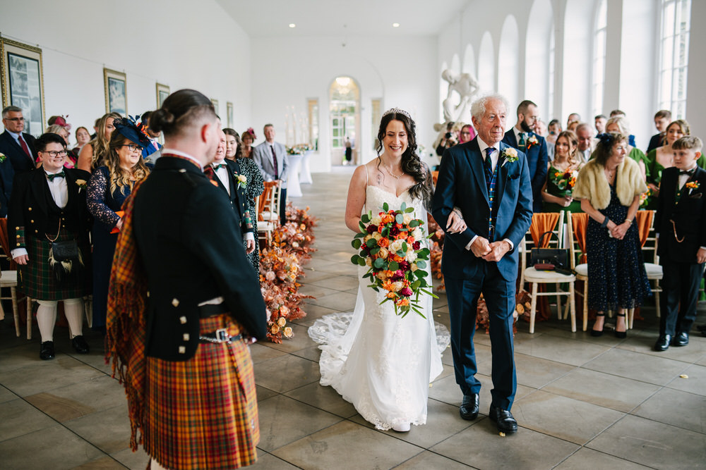 Bride walking the isle at Margam Orangery with burnt orange flowers and decor